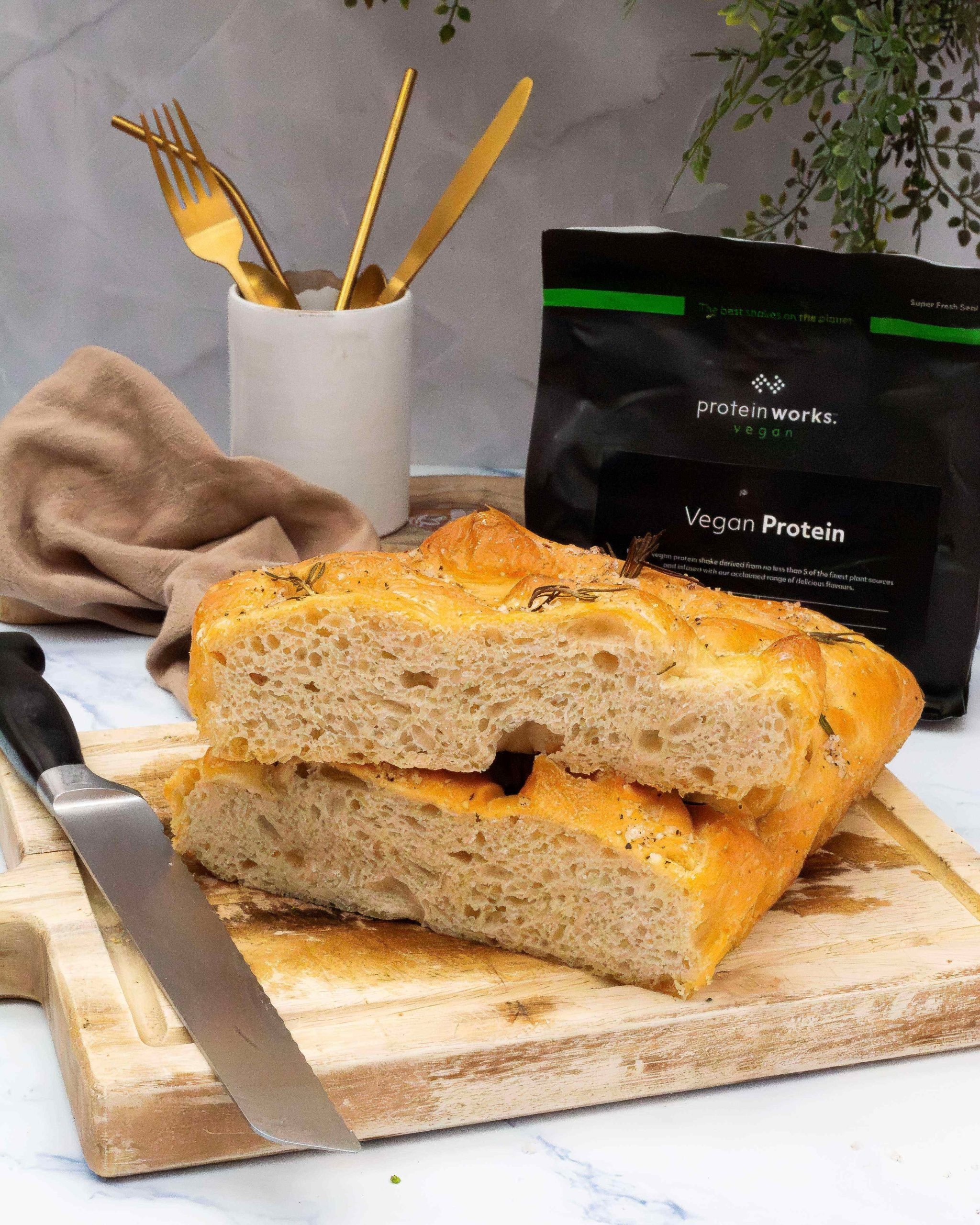 A slice of focaccia bread sitting on top of a loaf of focaccia. Vegan Protein pouch and gold cutlery in the background